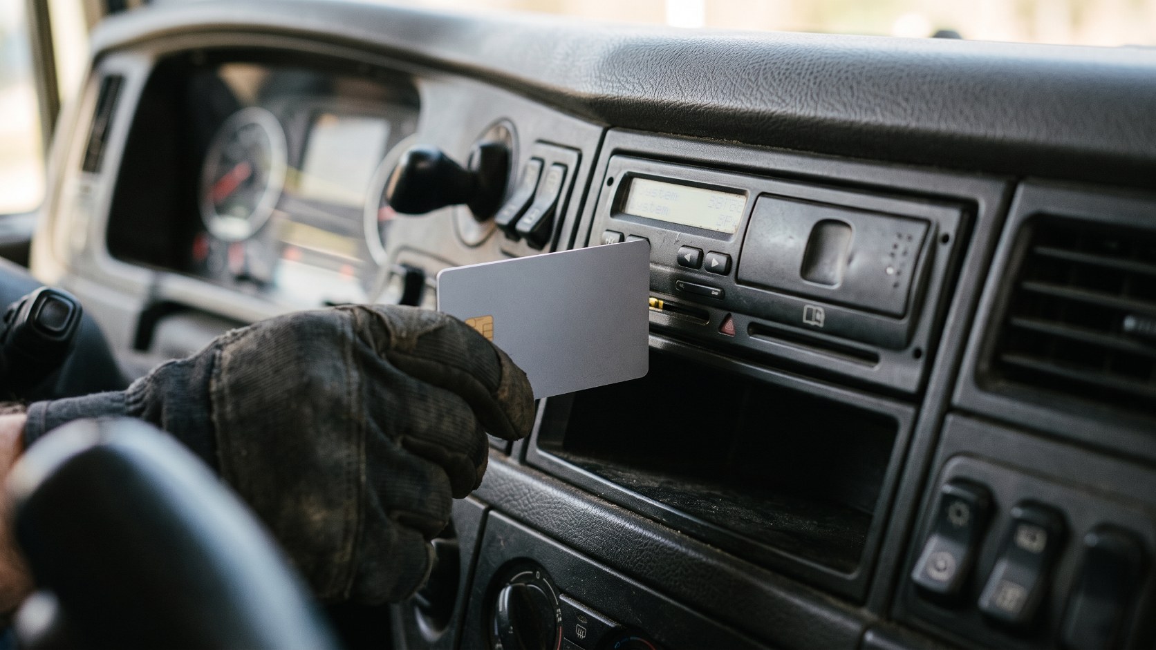 Gloved hand inserting a driver tachograph card into the digital tachograph unit slot on an HGV dashboard