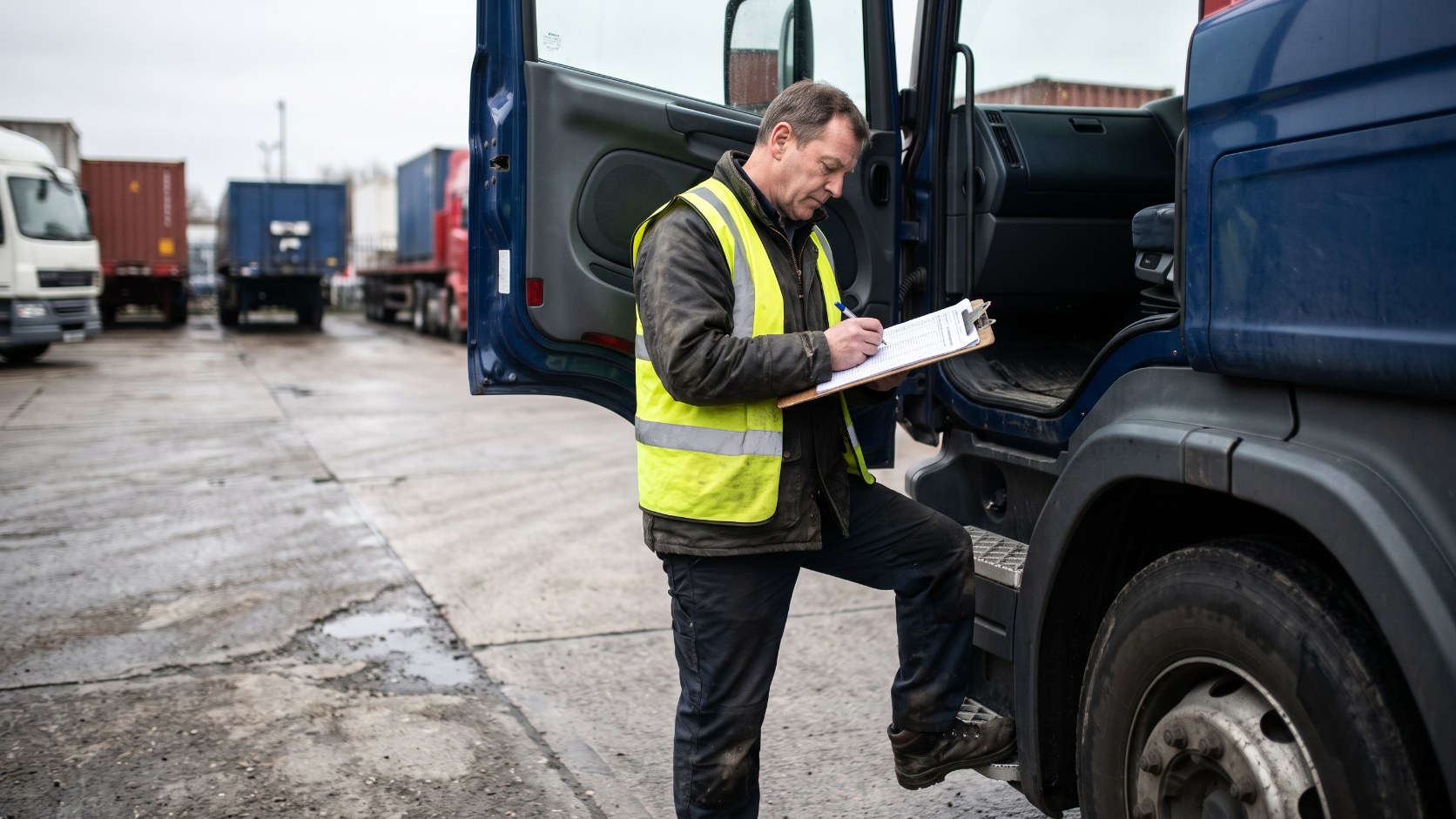 HGV driver filling in a paper form on a clipboard beside his lorry at a depot